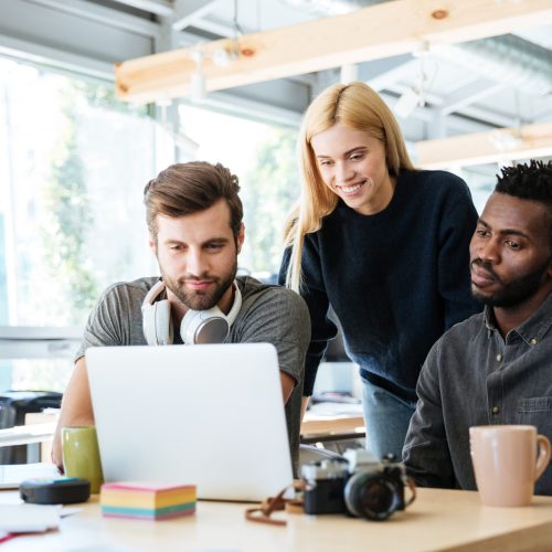 Photo of young smiling colleagues sitting in office coworking. Looking aside talking with each other.